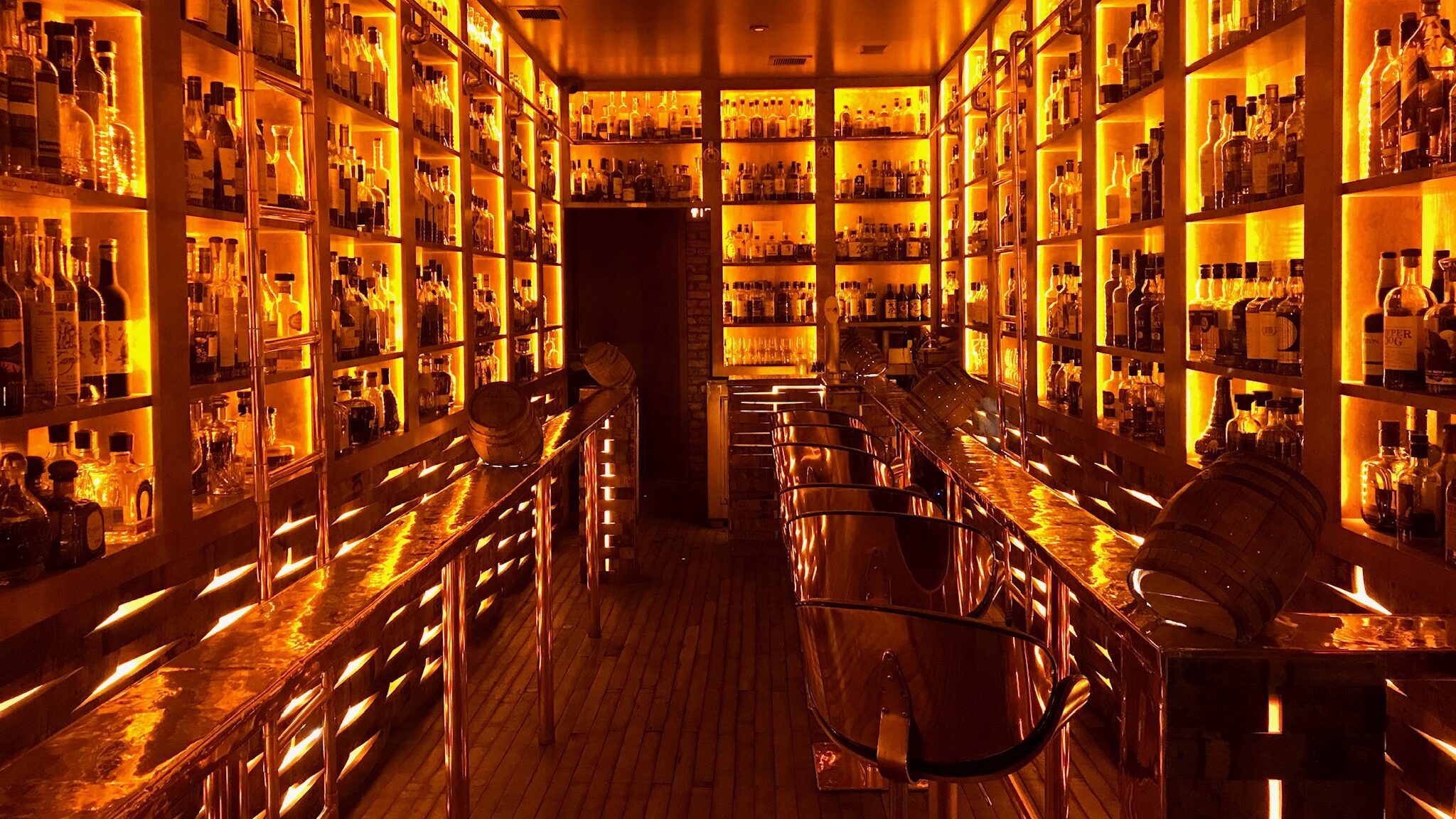 Black and white image of inside copper and oak bar with 8 seats and walls lined with spirits